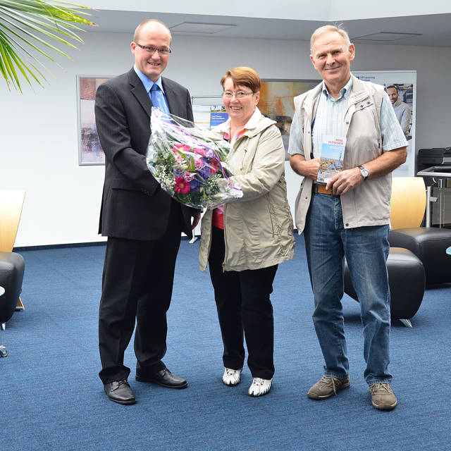 Filialleiter Thorsten Seiler überreicht den Blumengruß an Angela Schmitt. Ihr Mann Heinrich freut sich über den Reisegutschein. (Foto: Volksbank)